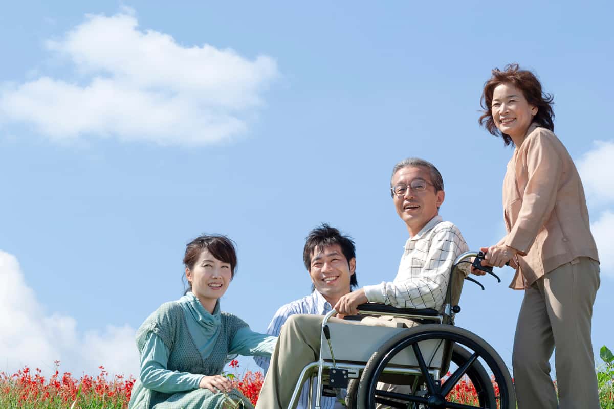 春の温かい青空の下で家族で花見をしている。車いすの父親をのせて話している。